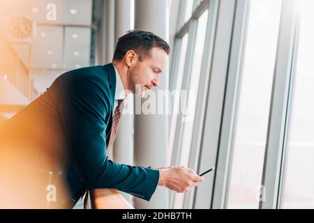Side view of mature businessman using mobile phone while leaning on railing in airport Stock Photo