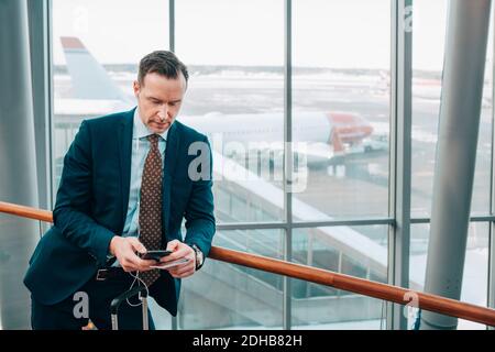 Businessman using mobile phone while leaning on railing in airport Stock Photo