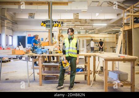 Full length portrait of confident young female trainee holding power tool while standing in workshop Stock Photo