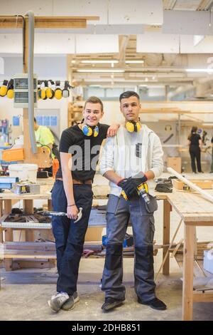 Full length of smiling young male trainee leaning on coworker's shoulder while holding work tools at workshop Stock Photo