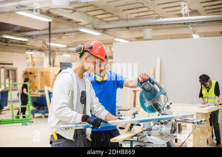 Teenage carpenter using level while standing by instructor with electric saw at illuminated workshop Stock Photo