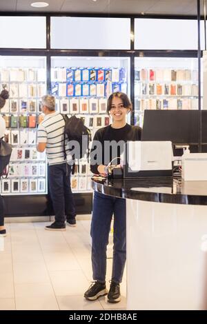 Full length portrait of confident female sales clerk standing by checkout against customers in shop Stock Photo