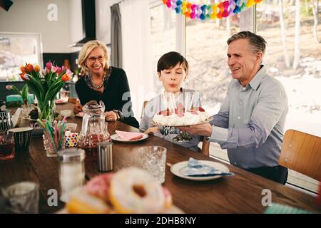 Boy blowing candles on birthday cake while sitting with grandparents at table Stock Photo