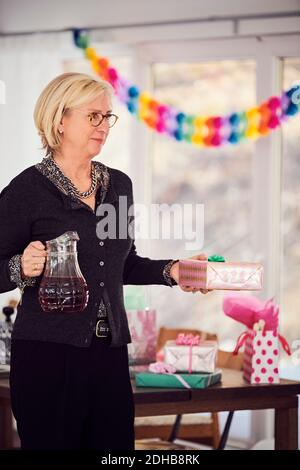 Senior woman holding gift box and juice while standing by table at home Stock Photo