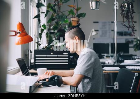 Side view of male hipster using laptop at desk in creative office Stock Photo