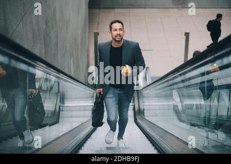 Confident businessman holding drink while moving on escalator in subway Stock Photo