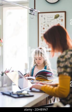 Side view of woman using laptop while sitting by girl studying at kitchen island Stock Photo