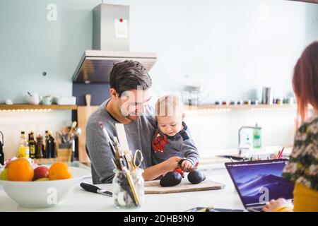 Smiling man carrying son holding tomato on cutting board at kitchen island Stock Photo