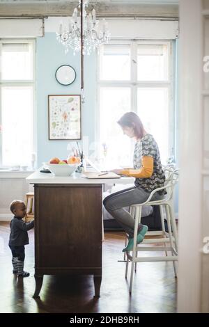 Full length of woman working on laptop with son standing by kitchen island at home Stock Photo