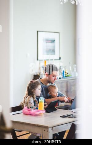 Father using laptop while sitting with cute children at table Stock Photo