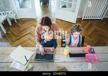 High angle view of mother working on laptop while sitting with cute son by daughter using digital tablet at table Stock Photo