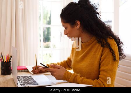 Mixed race woman using smartphone and laptop sitting at desk writing Stock Photo