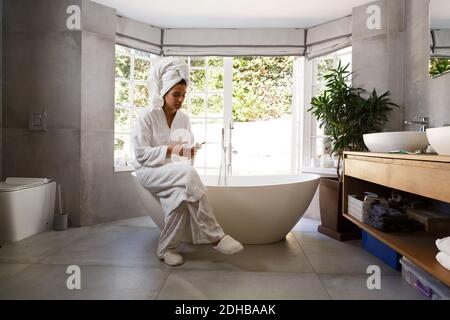 Mixed race woman wearing bathrobe sitting on bathtub using smartphone Stock Photo