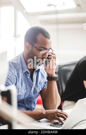 Businessman talking over smart phone while using laptop on desk in office Stock Photo