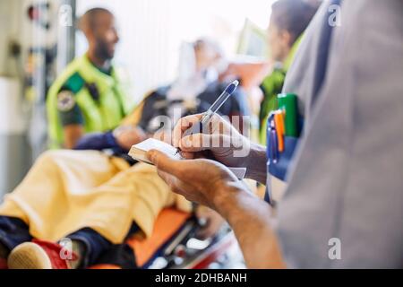 Close-up midsection of male nurse writing medical report against paramedics and patient Stock Photo