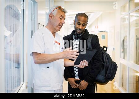 Senior doctor discussing with young man over digital tablet during routine check up in corridor Stock Photo