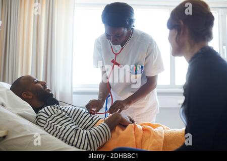 Female doctor examining patient by visitor during routine check up in hospital ward Stock Photo