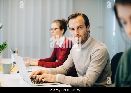 Portrait of confident businessman using laptop while sitting amidst female colleagues at creative office Stock Photo