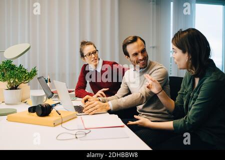 Smiling business coworkers discussing while sitting at desk with laptop in creative office Stock Photo