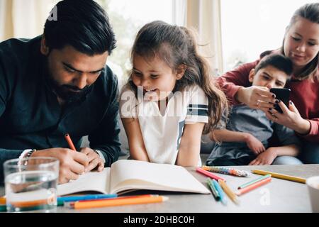 Father teaching drawing to daughter while woman using smart phone with autistic son in background Stock Photo