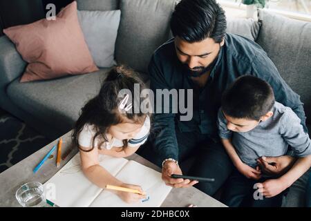 High angle view of father showing mobile phone to son while daughter drawing in book at home Stock Photo