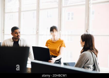 Female software engineer explaining to coworkers while sitting against window in office Stock Photo