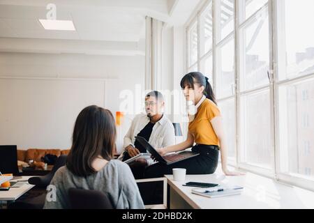 Female computer engineer discussing with colleagues while sitting on desk in office Stock Photo