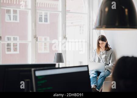 Focused young female programmer using laptop while sitting by window in creative office Stock Photo
