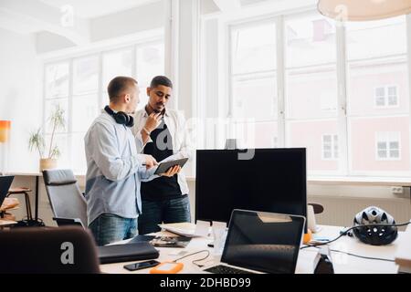 Male IT engineers discussing over digital tablet while standing against window in creative office Stock Photo