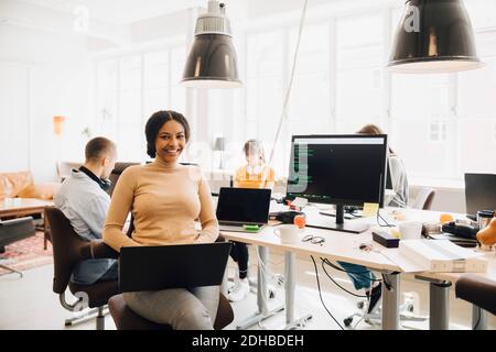 Portrait of smiling female programmer using laptop while coworkers working in background Stock Photo