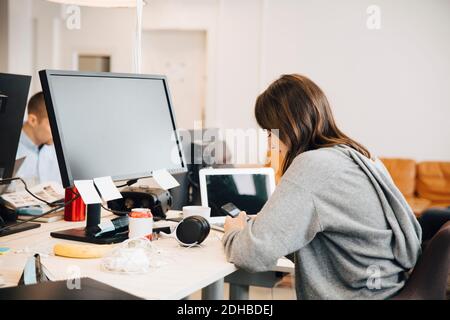 Side view of female programmer using mobile phone on desk while sitting in office Stock Photo