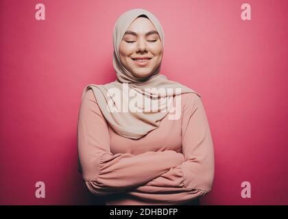Smiling Muslim woman with eyes closed standing against pink background Stock Photo