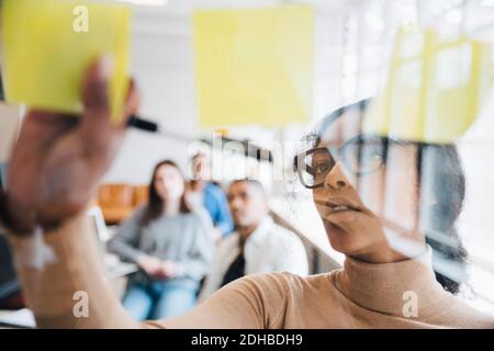 Female computer hacker sticking adhesive notes on glass with colleagues sitting in background Stock Photo