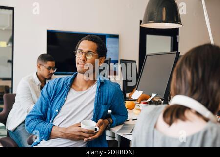 Thoughtful male computer programmer looking away while sitting by colleagues at desk in office Stock Photo