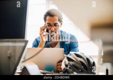 Computer programmer talking on smart phone while using laptop in creative office Stock Photo