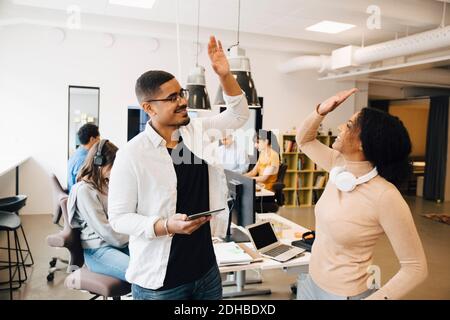 Smiling programmers doing high-five while standing with colleagues working in background Stock Photo