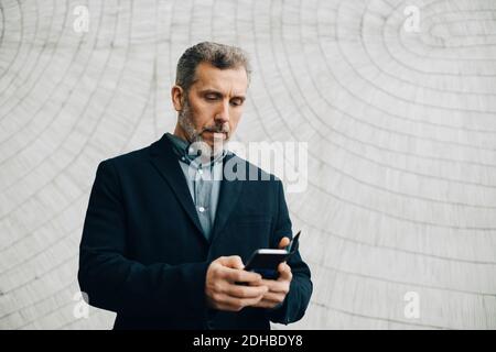 Mature businessman using smart phone against wall at office Stock Photo