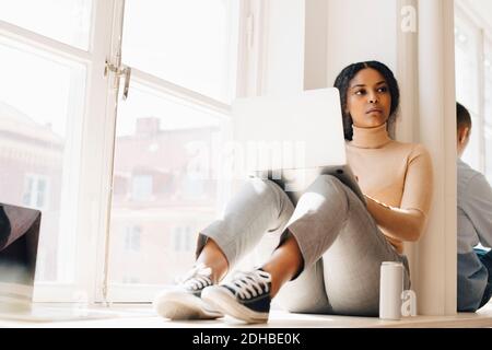 Full length of female computer programmer looking away while sitting by window in office Stock Photo
