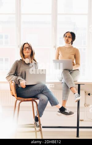 Portrait of female computer programmers with laptops sitting at desk in office Stock Photo