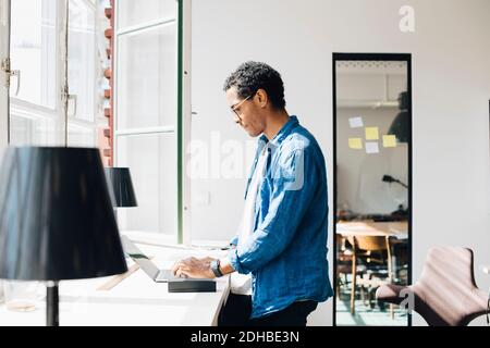 Side view of programmer working on laptop at desk by window in office Stock Photo