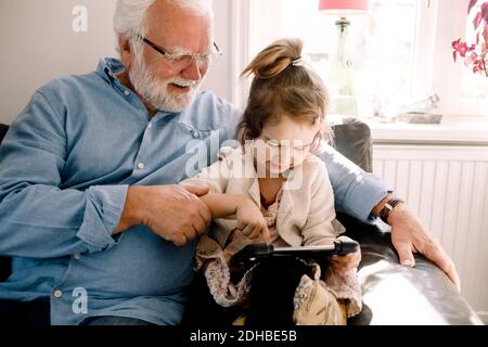Girl using digital tablet while sitting with grandfather on sofa at home Stock Photo