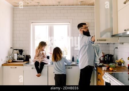 Girl looking at working sister while father doing chores in kitchen Stock Photo