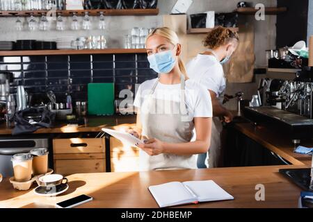 blonde waiter in medical mask holding digital tablet near notebook, coffee to go, and barista working on background Stock Photo
