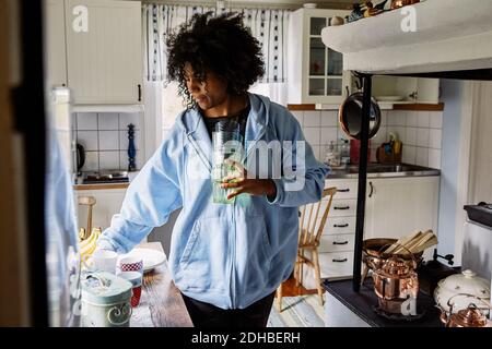 Young woman with curly hair holding drinking glasses while standing in kitchen at home Stock Photo
