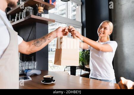 smiling blonde woman taking paper bag from tattooed barista on blurred foreground Stock Photo