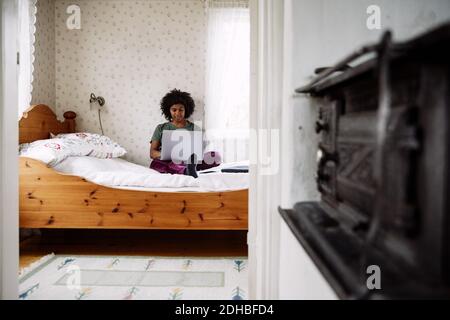 Young woman using laptop while relaxing on bed at home seen through doorway Stock Photo