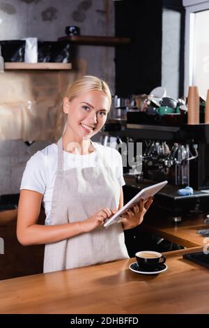 smiling blonde barista using digital tablet near cup of coffee on bar counter Stock Photo