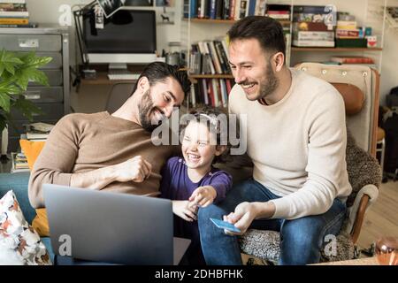 Happy fathers with daughter shopping online on laptop through credit card while sitting on chairs at home Stock Photo