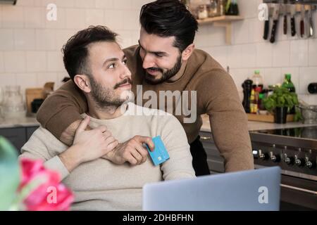 Romantic gay couple doing online shopping through laptop in kitchen at home Stock Photo