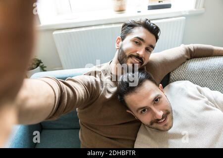 Smiling gay couple taking selfie while relaxing on sofa in living room at home Stock Photo